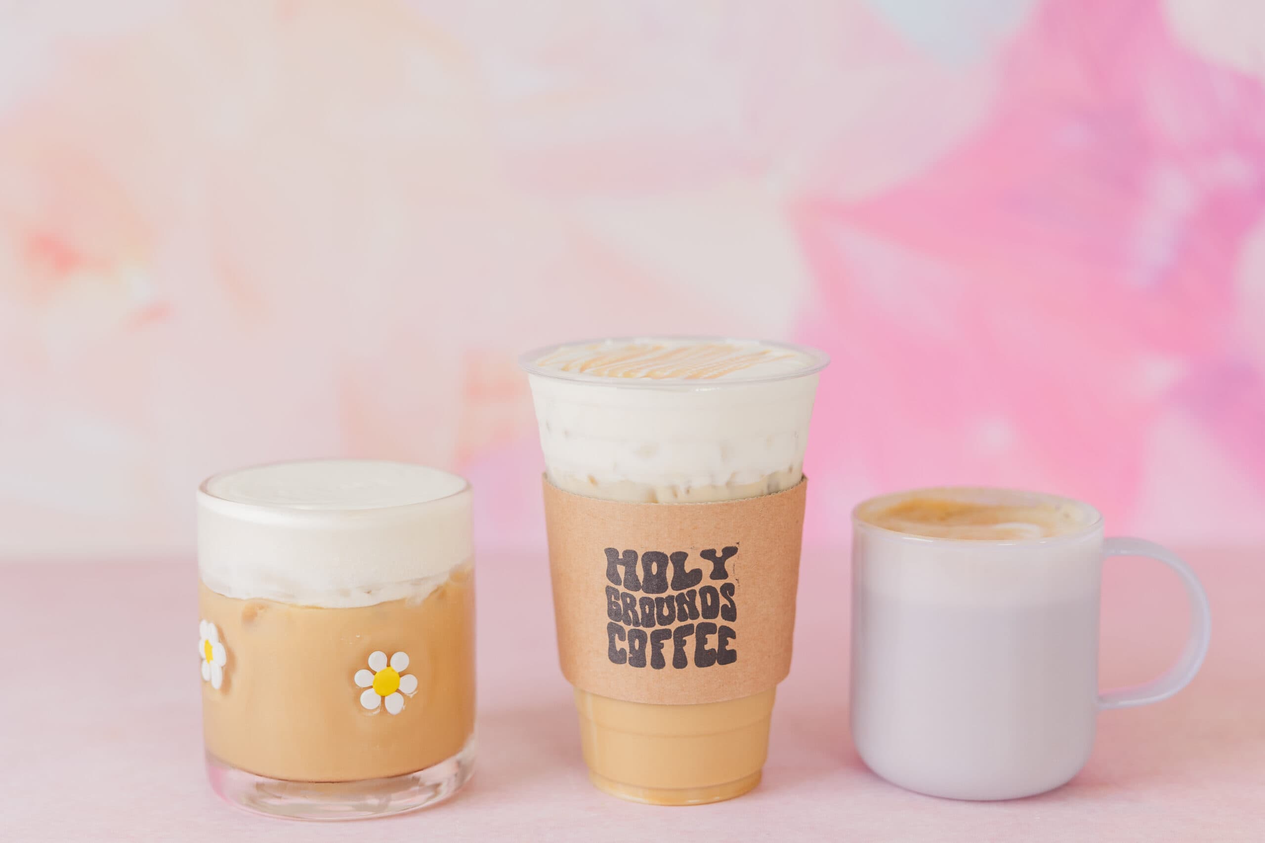 Three beautifully crafted coffee drinks from Holy Grounds Coffee, set against a soft pink and pastel background. The drinks include a caramel-drizzled iced coffee in a branded cup sleeve, a creamy iced latte in a glass with cute white daisies, and a warm, frothy latte in a translucent mug. A bright and inviting setup showcasing the spring seasonal menu.
