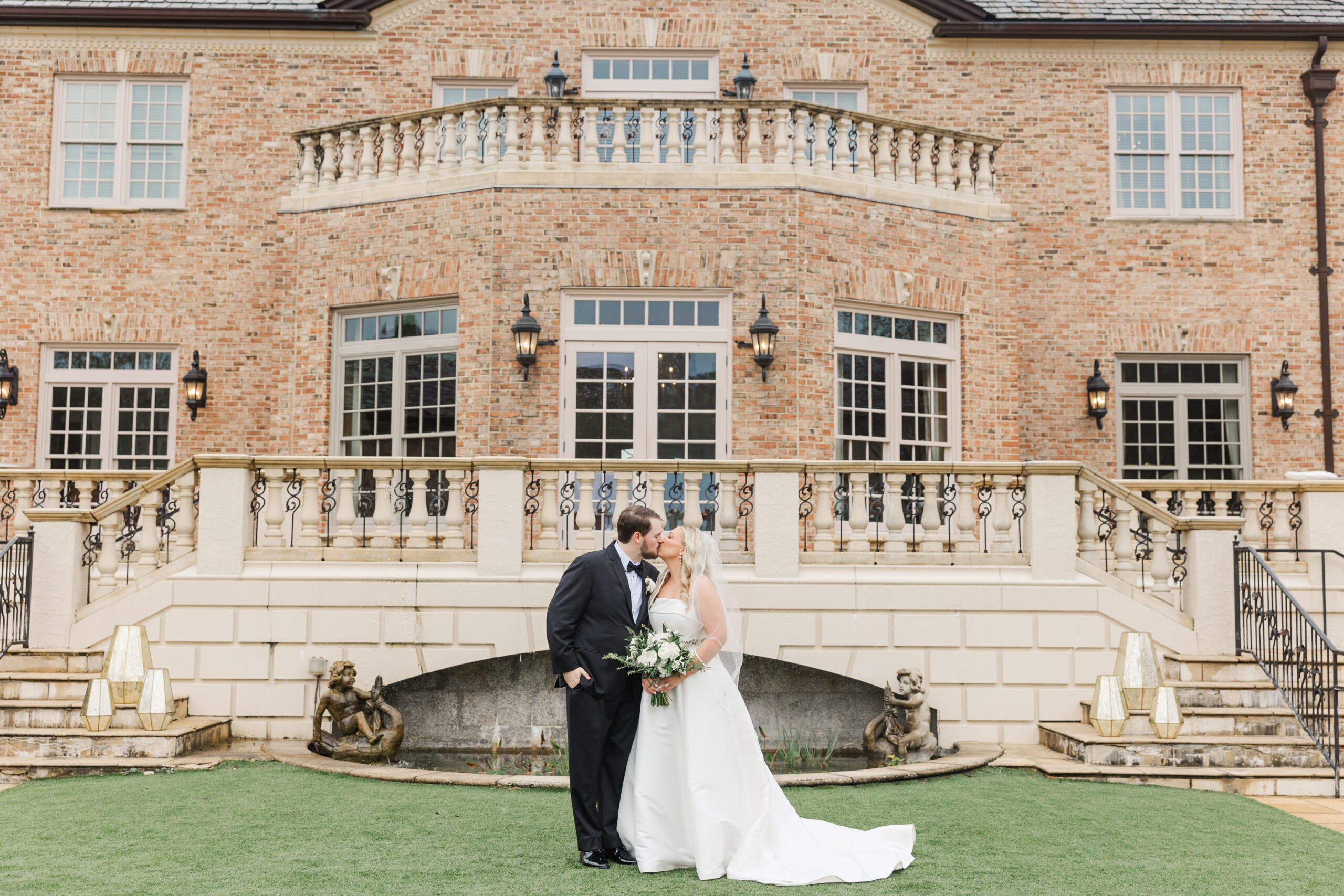 A newlywed couple shares a kiss in front of the grand Fountainview Mansion in Auburn, Alabama. The bride wears a classic white gown with a long train, and the groom is dressed in a black tuxedo. The mansion's elegant brick facade, intricate railings, and lush landscaping create a romantic and timeless wedding setting.