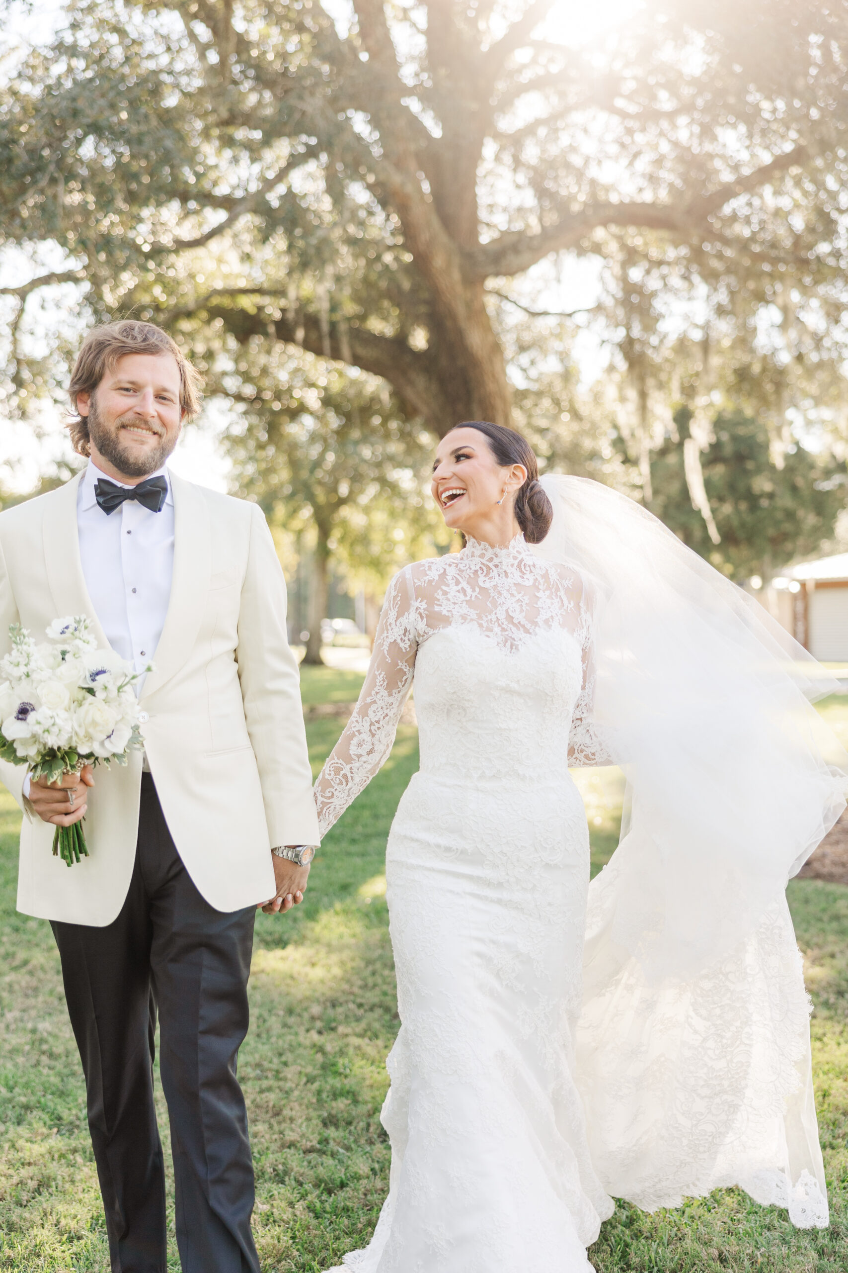 Bride and groom walking hand in hand under a large oak tree at their outdoor wedding in Orange Beach, Alabama. The bride, wearing a lace high-neck wedding gown with a flowing veil, smiles joyfully at her groom, who is dressed in a white tuxedo jacket and black bow tie, holding a bouquet of white flowers. Sunlight filters through the trees, creating a dreamy and romantic atmosphere.