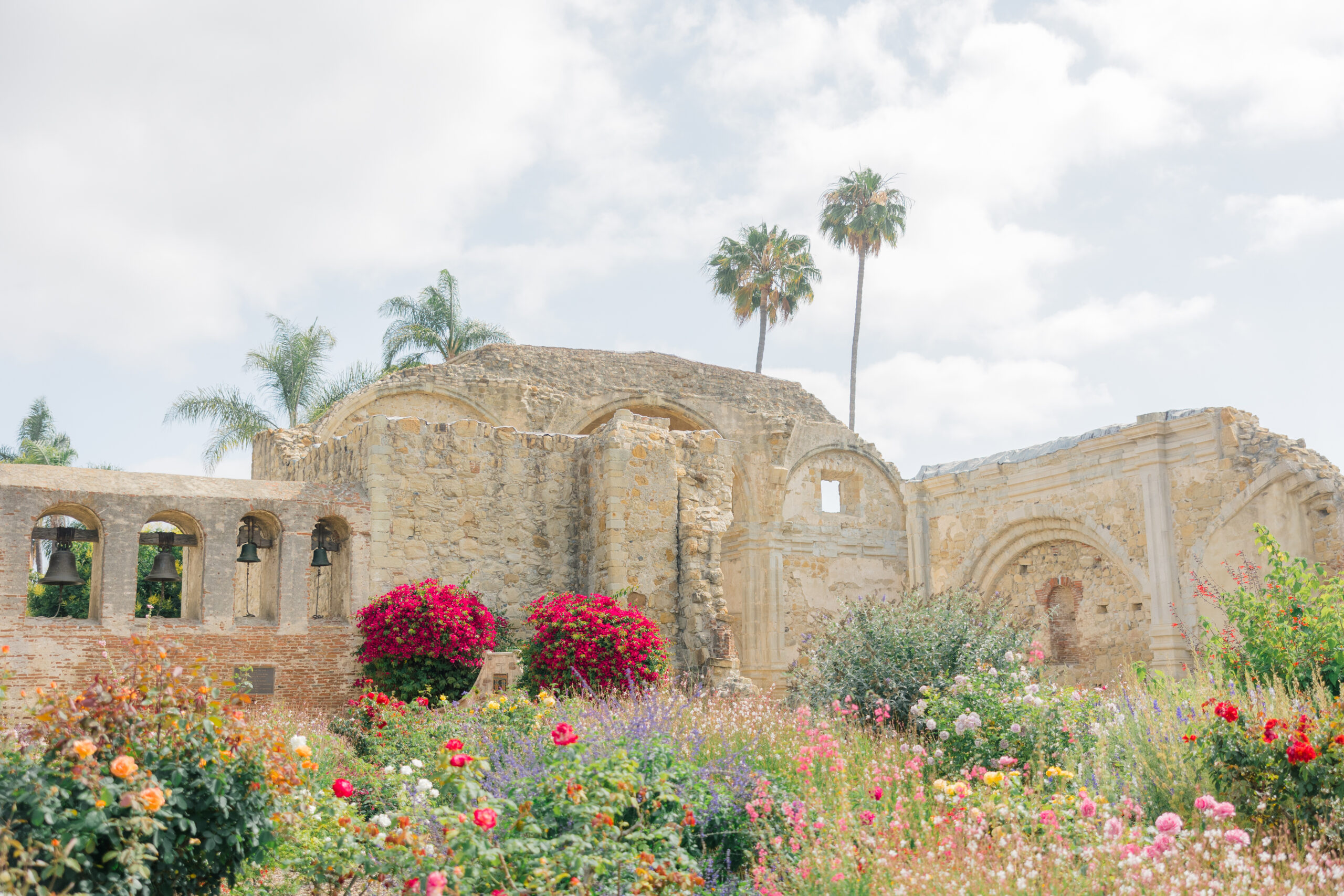 Historic stone ruins at Mission San Juan Capistrano surrounded by vibrant spring flowers, palm trees, and bell arches under a partly cloudy sky.