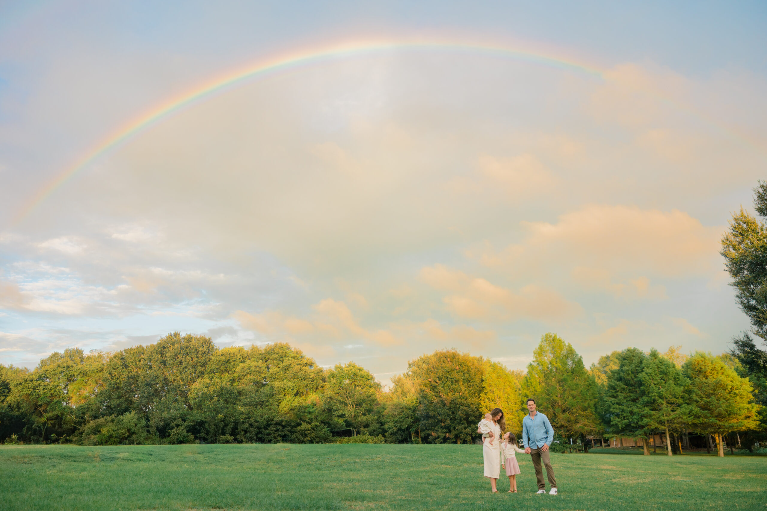 Family standing together in an open field at Colony Nature Park in Fairhope, Alabama as a full rainbow stretches across the sky after a light rain.